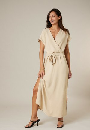 Femme souriante, portant une robe portefeuille beige à manches courtes, taille nouée, fente sur le côté, et des sandales à talons noires, debout sur un fond neutre.