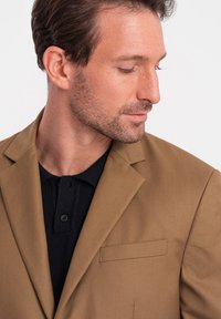 Ruskea bleiseri, jossa on viistot kaulukset, yksi rintatasku ja sileä pinta. Yllä tummanvärinen neulottu poolopaita.