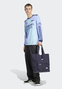 Blå gradient atletisk trøje med lange ærmer. Sort corduroy tote taske med forlommer, der har logo og mørkeblå detaljer. Sorte bukser og sko.
