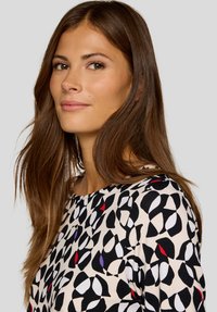 Patterned blouse featuring a cream background with black, red, and purple abstract shapes. Smooth texture and round neckline.