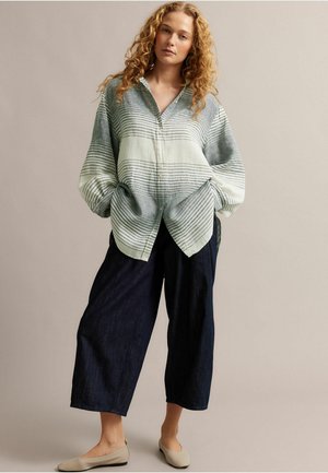 Kobieta o kręconych włosach, ubrana w luźną, w paski koszulę zapinaną na guziki, szerokie ciemne spodnie i beżowe baleriny, stojąca z rękami w kieszeniach.