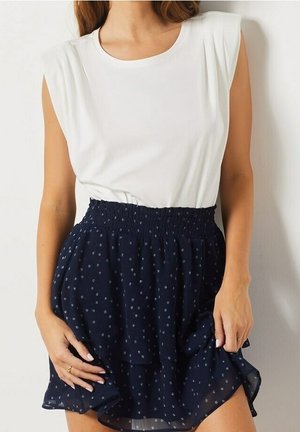 Nainen pukeutunut valkoiseen hihattomaan toppiin ja tummansiniseen, korkeavyötäröiseen hameeseen, jossa on pieniä valkoisia kukkakuoseja, pitää kiinni hameen helmasta.