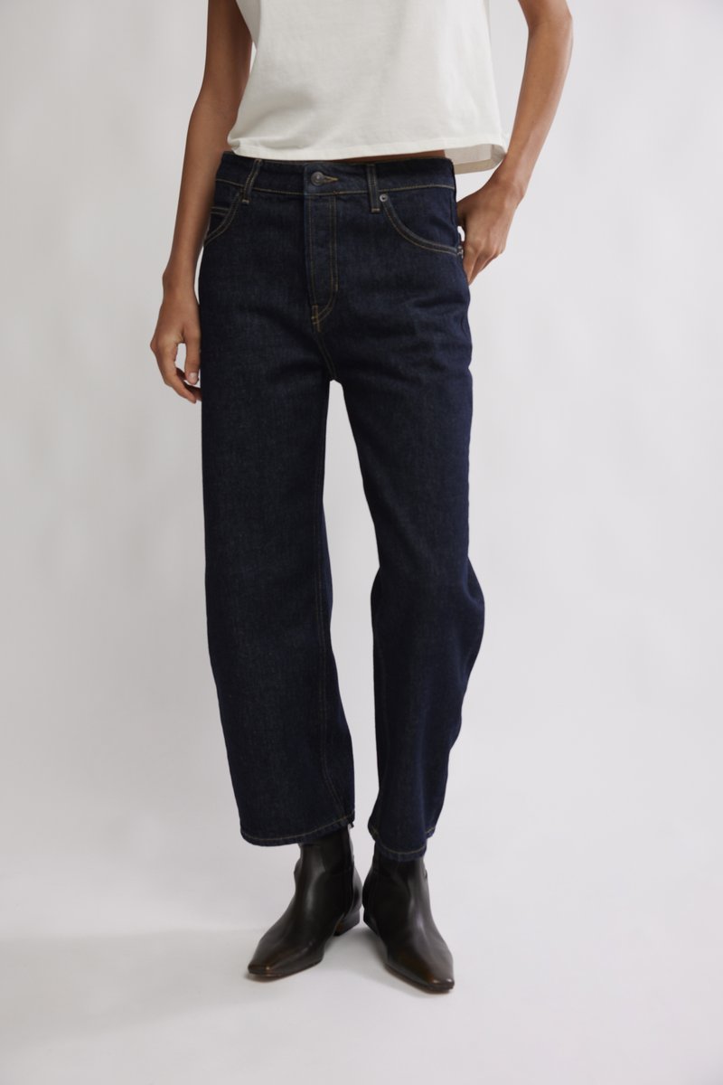 Jean en denim bleu foncé avec une coupe droite, coutures jaunes, fermeture par bouton et poches avant, associé à des bottines noires.