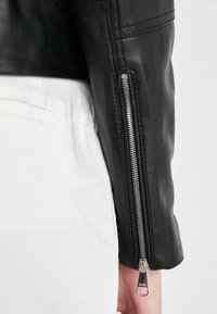 Ärmen på en svart läderjacka med en silverfärgad dragkedja och en jämn textur, med detaljerad sömnad längs axelsömmarna.