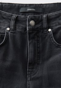 Close-up van de taille van zwarte spijkerbroek met zilveren knoop en klinknagels, voorzakken, riemlussen en het label van het merk "someday." aan de binnenkant van de kraag.