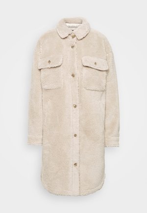 Béžový kabát z umělé kožešiny s límcem, dvěma náprsními kapsami a zapínáním na knoflíky vpředu. Má jemnou texturu a zaoblený spodní lem.