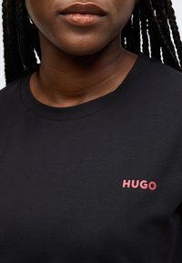 Fekete pamut rövid ujjú póló bordázott nyakkivágással, bal oldalon kis piros "HUGO" logóval. Sima textúra.