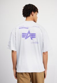 Lys grå oversized t-shirt med et lilla grafisk design på ryggen, der viser bogstaverne "A" samt trykt tekst ovenfor og nedenfor grafikken.