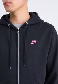 Juodas džemperis su pilnu užtrauktuku, su rožiniu Nike logotipu kairėje krūtinės pusėje. Pagamintas iš švelnios medžiagos, su raišteliais ir priekinių kišenių.