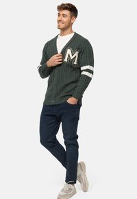 Groene kabelgebreide cardigan met witte strepen en een grote letter "M" op de borst, gecombineerd met donkere jeans en lichte sneakers.