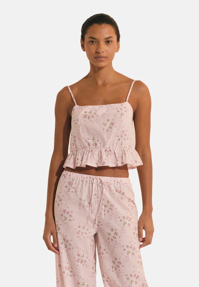 Mujer llevando un top de tirantes finos con estampado floral en rosa claro con dobladillo de volantes y pantalones sueltos a juego, sobre un fondo blanco.