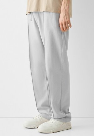 Person iført lysegrå løstsiddende bukser, beige skjorte og hvide sneakers, stående mod en ensfarvet hvid baggrund.
