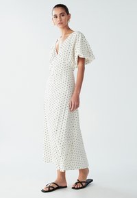 White polka dot dress with a deep V-neck and short flutter sleeves, featuring a flowing silhouette. Paired with black flat sandals.