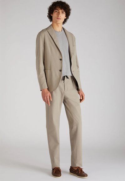 Blazer beige con tessuto testurizzato, a un bottone, due tasche frontali, abbinato a una t-shirt grigio chiaro e pantaloni beige, completato da scarpe marroni.
