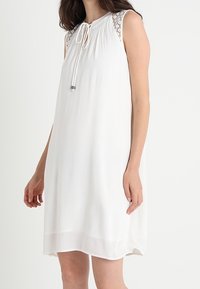 Biała bezrękawnikowa sukienka z zebranym dekoltem, dekoracyjnym koronkowym wykończeniem na ramionach oraz wiązaniem z przodu. Gładki, lekki materiał.