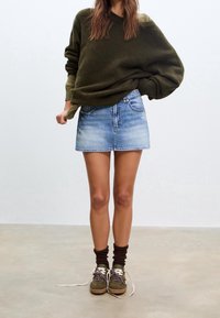 Pull oversize vert olive, mini-jupe en jean bleu clair, chaussettes marron, et chaussures vertes/marron avec lacets à motifs sur un fond neutre.