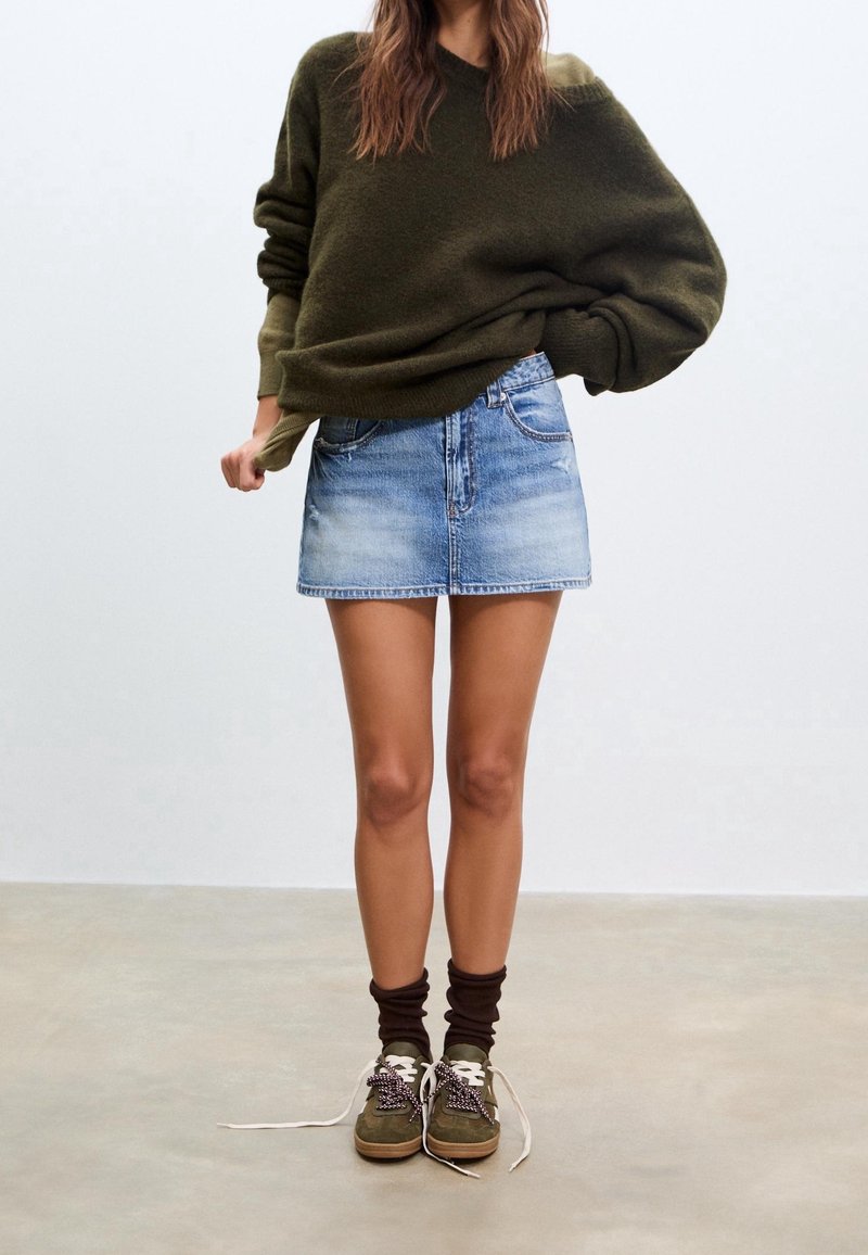 Pull oversize vert olive, mini-jupe en jean bleu clair, chaussettes marron, et chaussures vertes/marron avec lacets à motifs sur un fond neutre.