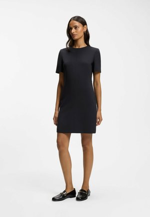 Femme portant une robe noire simple à manches courtes, longueur genou, et des mocassins noirs, debout devant un fond blanc uni.