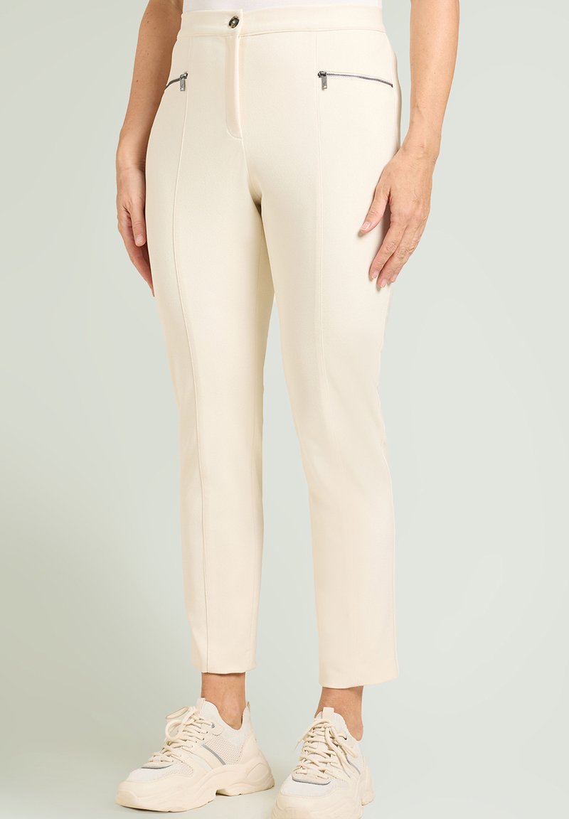 Roomkleurige, op maat gemaakte broek met ritszakken aan de voorkant, slanke pasvorm en gladde textuur, gecombineerd met lichtbeige sportieve sneakers.