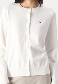 Weißer Cardigan aus weichem Strickmaterial mit rundem Ausschnitt, Knopfverschluss, gerippten Bündchen und kleinem Logo auf der linken Brust.