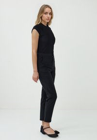 Une jeune femme se tient de profil, portant un haut noir ajusté et un pantalon noir, avec des ballerines, devant un fond blanc uni.