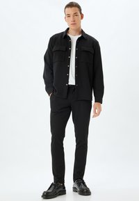 Camicia nera a maniche lunghe con due tasche frontali, chiusura con bottoni e t-shirt bianca sotto, abbinata a pantaloni neri slim-fit e scarpe nere.