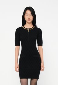 Femme portant une robe noire ajustée avec des manches jusqu'au coude et un détail de chaîne dorée au col, se tenant devant un fond blanc.