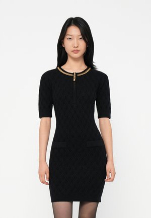 Woman wearing a fitted black textured dress with elbow-length sleeves and a gold chain detail at the neckline, standing against a white background.