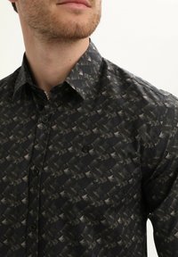 Camisa negra de botones con un patrón geométrico en tonos oscuros y apagados, con un logo sutil en el lado izquierdo del pecho y una textura suave.