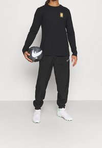 Schwarzes Langarmshirt mit kleinem Grafikdruck, schwarze Jogginghosen mit seitlichem Logo, hält einen schwarz-weißen strukturierten Fußball und weiße Fußballschuhe.