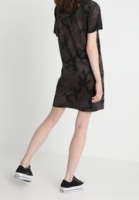 Black camouflage short-sleeve dress with a loose silhouette and a round neckline, worn with black sneakers, shown from a side angle.