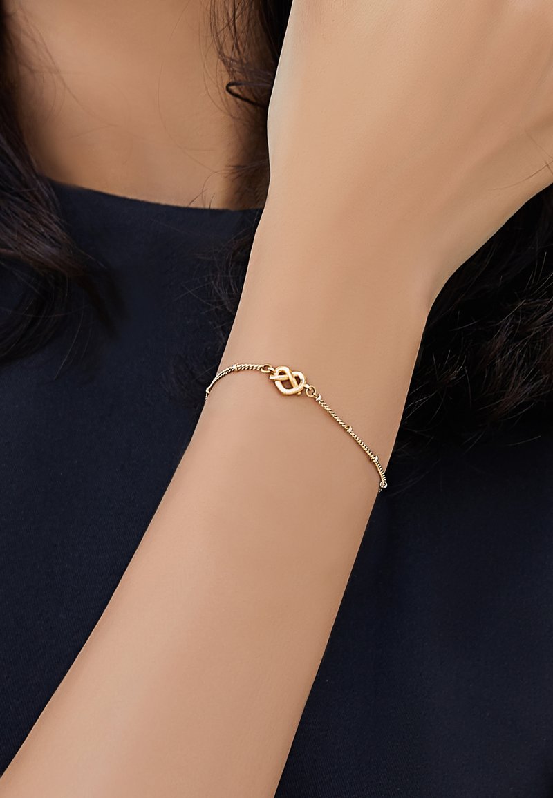 Bracciale in tono oro con un design a nodo attorcigliato, con una finitura liscia sui collegamenti a catena, indossato su un polso su uno sfondo scuro.