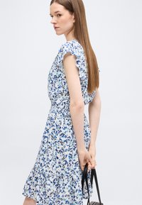 Γυναίκα με μακριά ίσια μαλλιά που φοράει αμάνικο μπλε και λευκό φόρεμα με λουλούδια, κρατώντας μια μαύρη τσάντα πίσω από την πλάτη της.