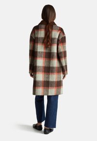Wollen geruite jas in bruin, crème en oranje, met een bruine kraag. Losse pasvorm, middellang, gecombineerd met blauwe denim broeken en zwarte schoenen.
