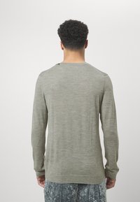 Langärmliger, olivgrüner Pullover mit glatter Textur und rundem Ausschnitt. Verfügt über einen Knopfdetails an der Schulter. Getragen mit gemusterten grauen Hosen.