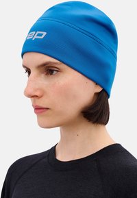 Jonge persoon die een felblauwe muts met wit logo draagt, een zwart shirt, naar links kijkt, met kort donker haar zichtbaar onder de muts.