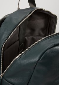 Sac à dos en cuir vert foncé avec une fermeture éclair argentée ouverte, montrant un intérieur en tissu noir vide et une sangle noire à l'intérieur.