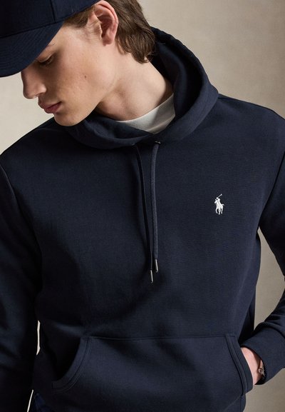 Jonge man draagt een marineblauwe hoodie met een wit polo-spelerlogo en een bijpassende marineblauwe pet, kijkt naar beneden met één hand in zijn zak.
