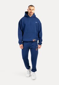 Marineblauer Hoodie mit großem Fronttasche und gesticktem Logo. Kombiniert mit passenden Jogginghosen mit Logo- und Etikettenakzenten. Weiße Sneakers.