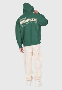 Personne portant un sweat à capuche vert DROPSIZE avec la capuche levée et un pantalon cargo beige, se tenant de dos contre un fond uni.
