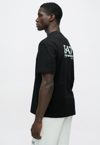 Férfi fekete EA7 Emporio Armani logós pólóban, fehér rövidnadrágban, profilból, sima fehér háttér előtt áll.