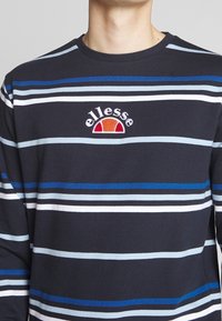 Persona che indossa una maglietta a maniche lunghe a righe nera, blu, bianca e grigia con un logo ellesse rosso e arancione sul petto.