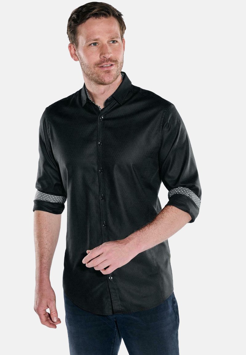 Mann in schwarzem Hemd mit auffälligen Ärmeln, das hochgekrempelt ist, kombiniert mit dunkelblauen Jeans, stehend vor einem schlichten Hintergrund.