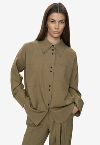 Camisa marrón de botón con textura, cuello grande, mangas largas y botones negros. Cuenta con un bolsillo en el pecho y un ajuste relajado.