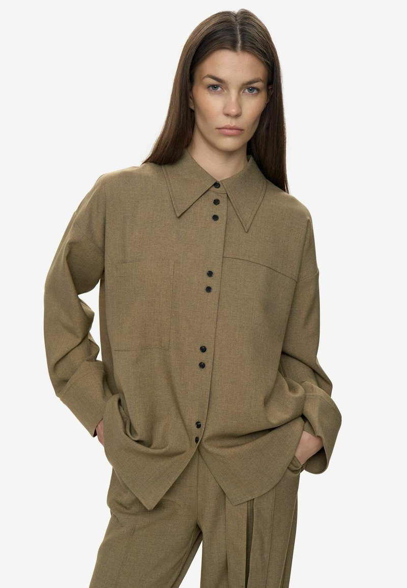 Camisa marrón de botón con textura, cuello grande, mangas largas y botones negros. Cuenta con un bolsillo en el pecho y un ajuste relajado.