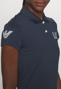 Polo din bumbac de culoare bleumarin, cu material texturat, croială slim fit, mâneci scurte și guler ascuțit. Accente cu logo pe piept și pe mânecă.