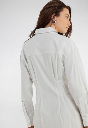 Mujer con cabello largo y castaño, vestida con un vestido camisa blanco de manga larga ajustado, de espaldas y mirando hacia un lado.