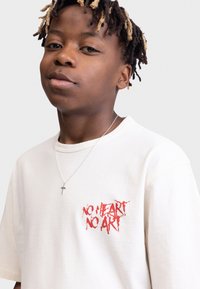 T-shirt en coton beige clair à manches courtes. Présente un texte graphique rouge "AUCUN COEUR AUCUN ART" sur le côté gauche de la poitrine. Collier en chaîne argentée avec une croix.