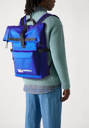 Zaino in nylon viola con parte superiore arrotolata, spallacci neri regolabili, tasca frontale e dettagli del logo bianco. Indossato con un maglione verde a coste.