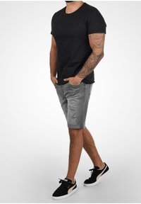 T-shirt noir à manches courtes, short gris délavé et baskets noires avec des accents crème. Le modèle est debout, les mains dans les poches.